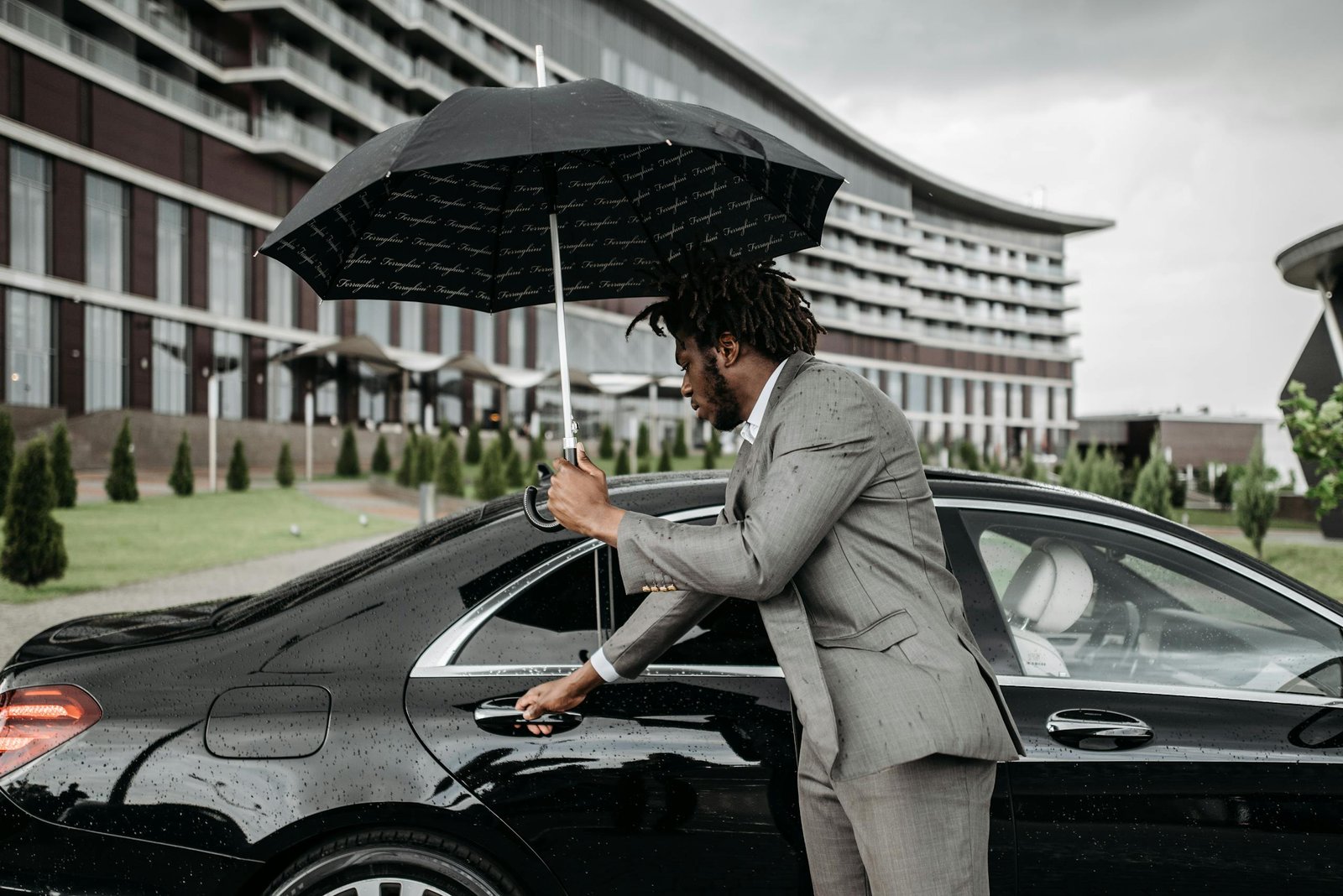 African American man in suit with umbrella opening car door outside modern building.